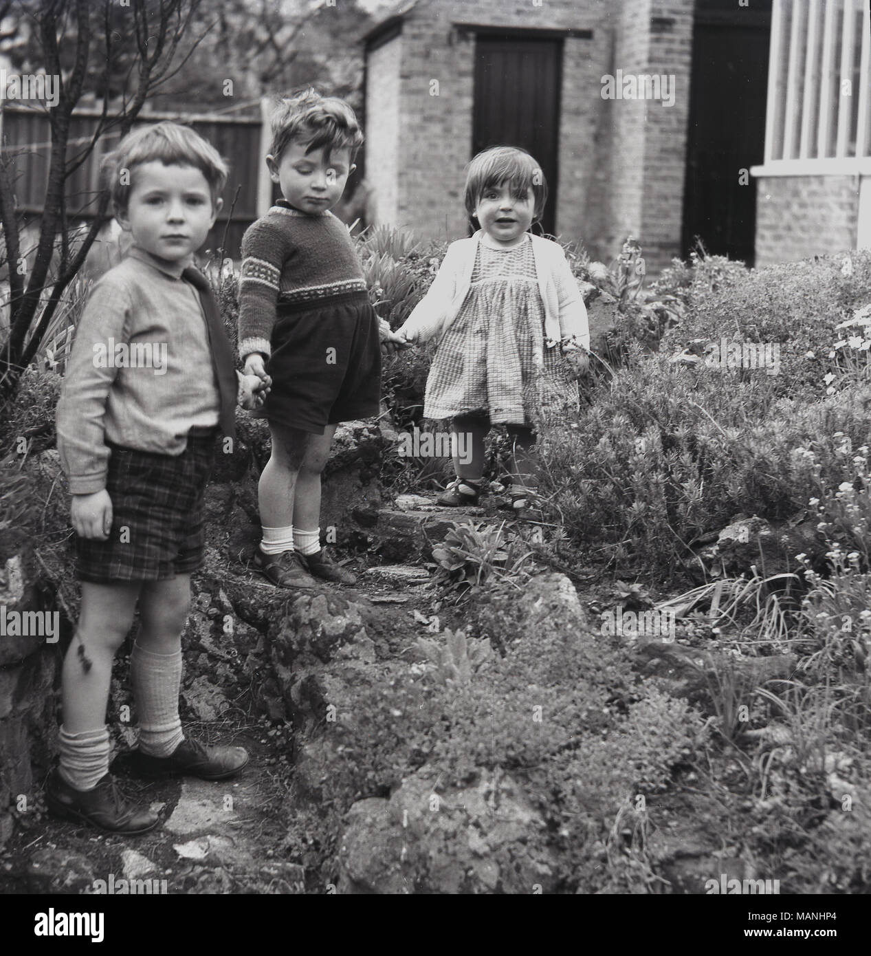 Années 1960, historiques, trois jeunes enfants jouer dehors par quelques pas dans un jardin de rocaille et tenir la main. Banque D'Images
