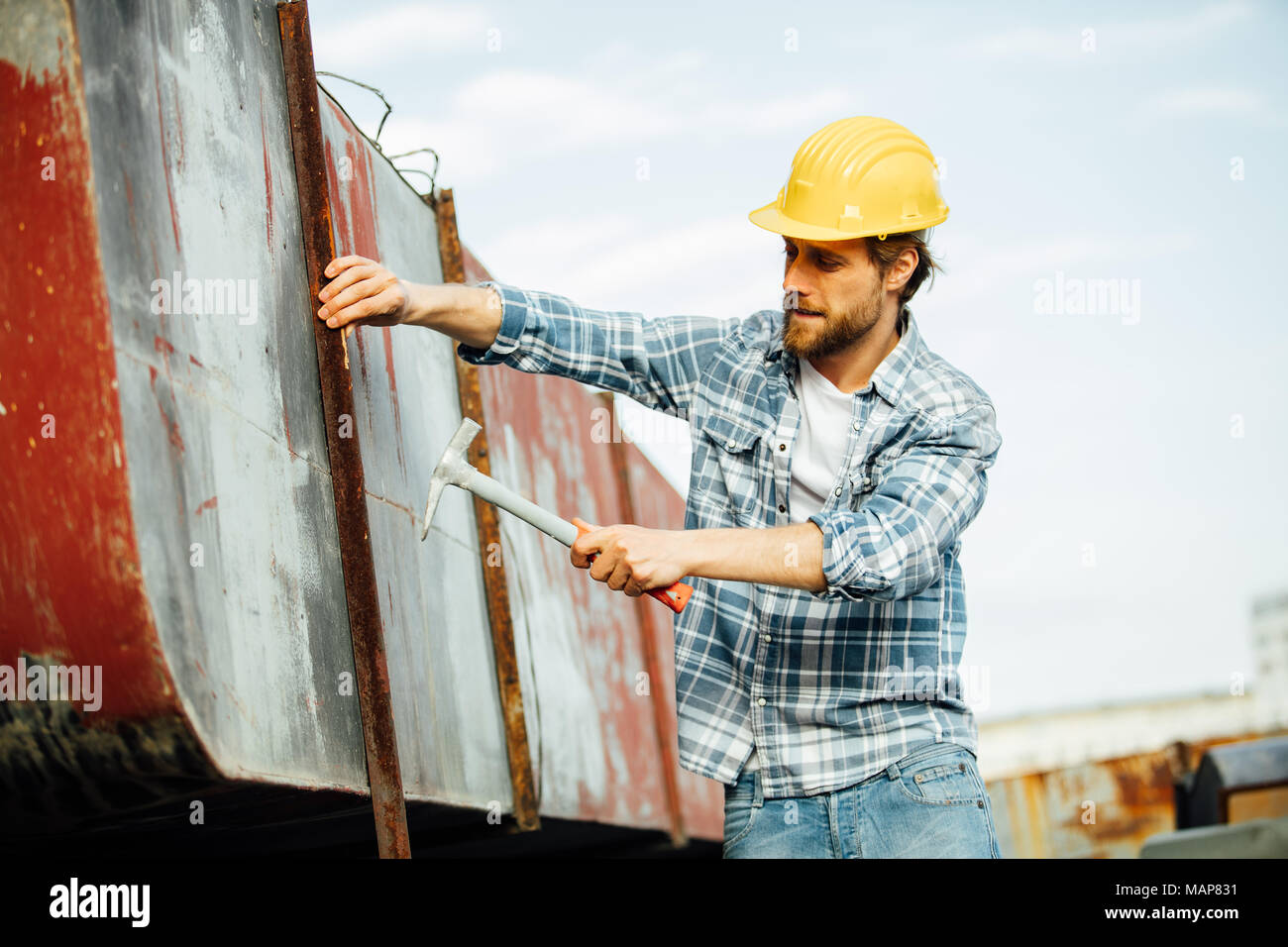 Homme fort avec barbe en chemise à carreaux, très concentré, martelage ou essayer de réparer certains objets rouillés qui exigent de l'attention, à l'extérieur sur un site Banque D'Images