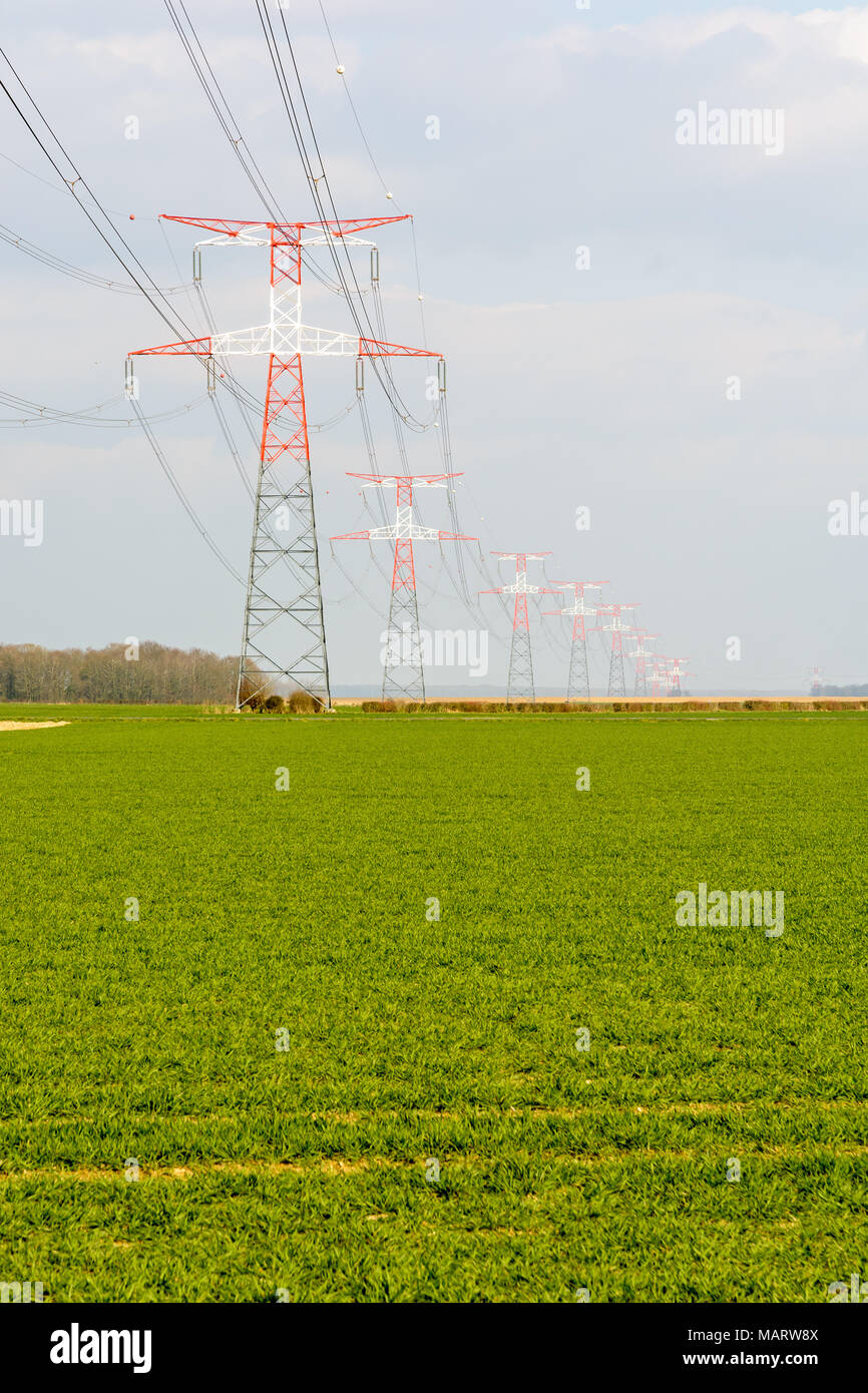 Une ligne électrique aérienne de tension soutenue par les tours de transmission rouge et blanche à travers la campagne française, avec un champ cultivé dans l'avant-plan. Banque D'Images