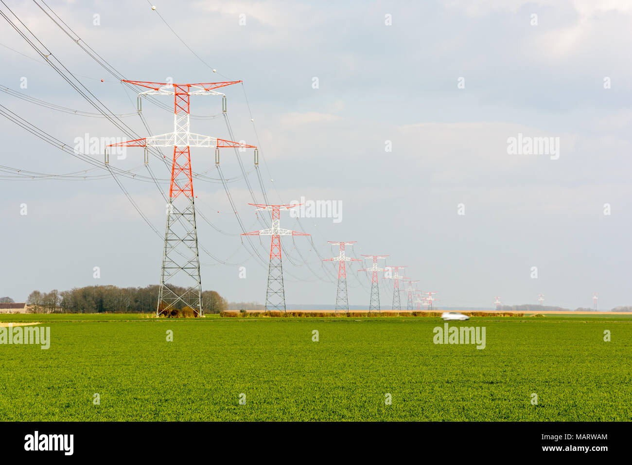 Une ligne électrique aérienne de tension soutenue par les tours de transmission rouge et blanc passant sur un champ cultivé et une route de campagne avec une voiture. Banque D'Images