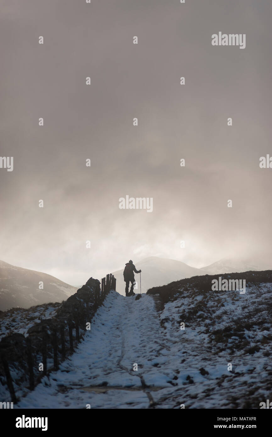 Un homme qui marche dans la neige avec l'aide d'un bâton de marche le long d'un chemin dans l'hiver sous un ciel sombre Banque D'Images