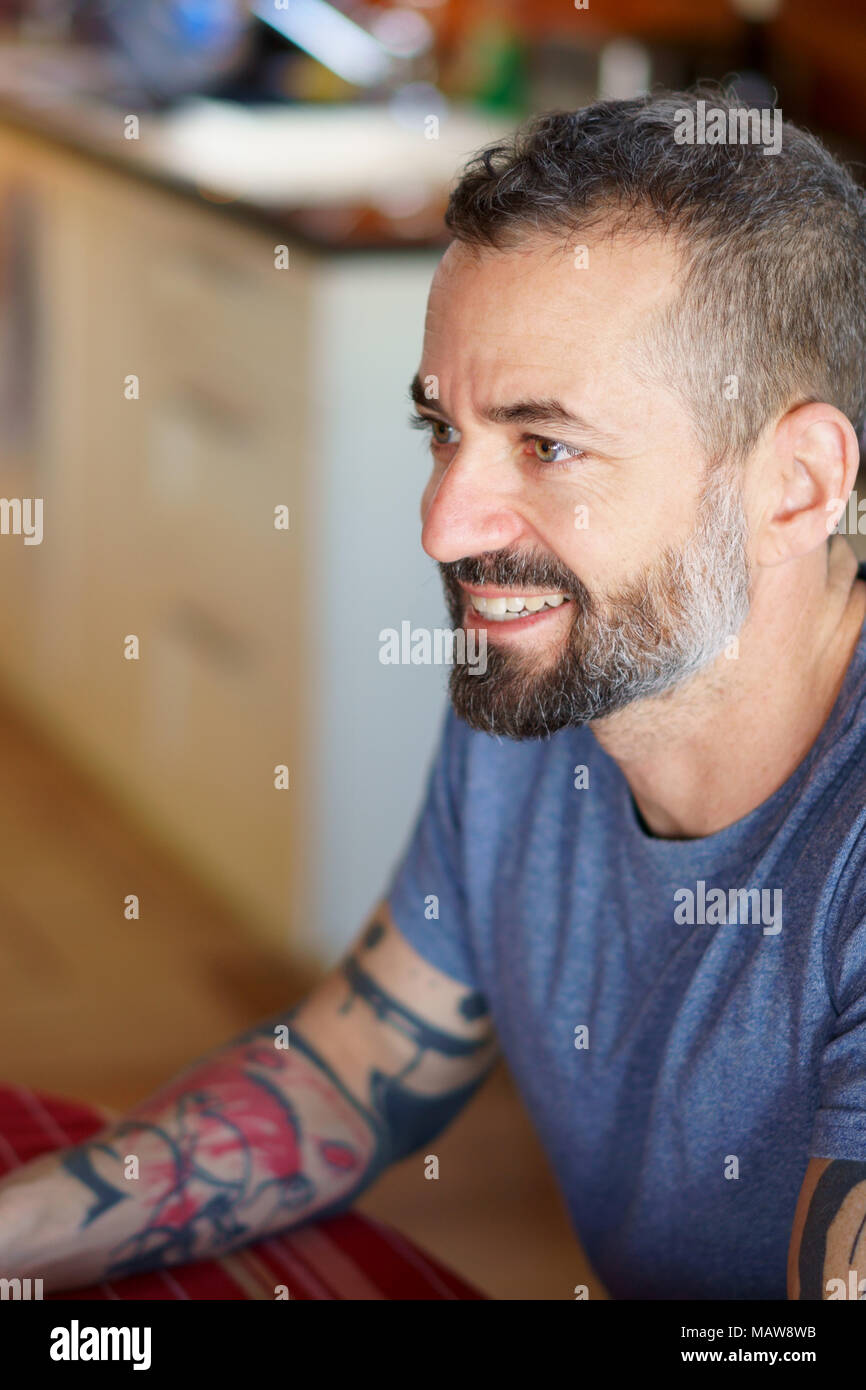 Le milieu de 30 ans barbe et tatoué homme souriant à la table de la cuisine avec une nappe rouge et portant un tee-shirt bleu et une grande attention. Banque D'Images