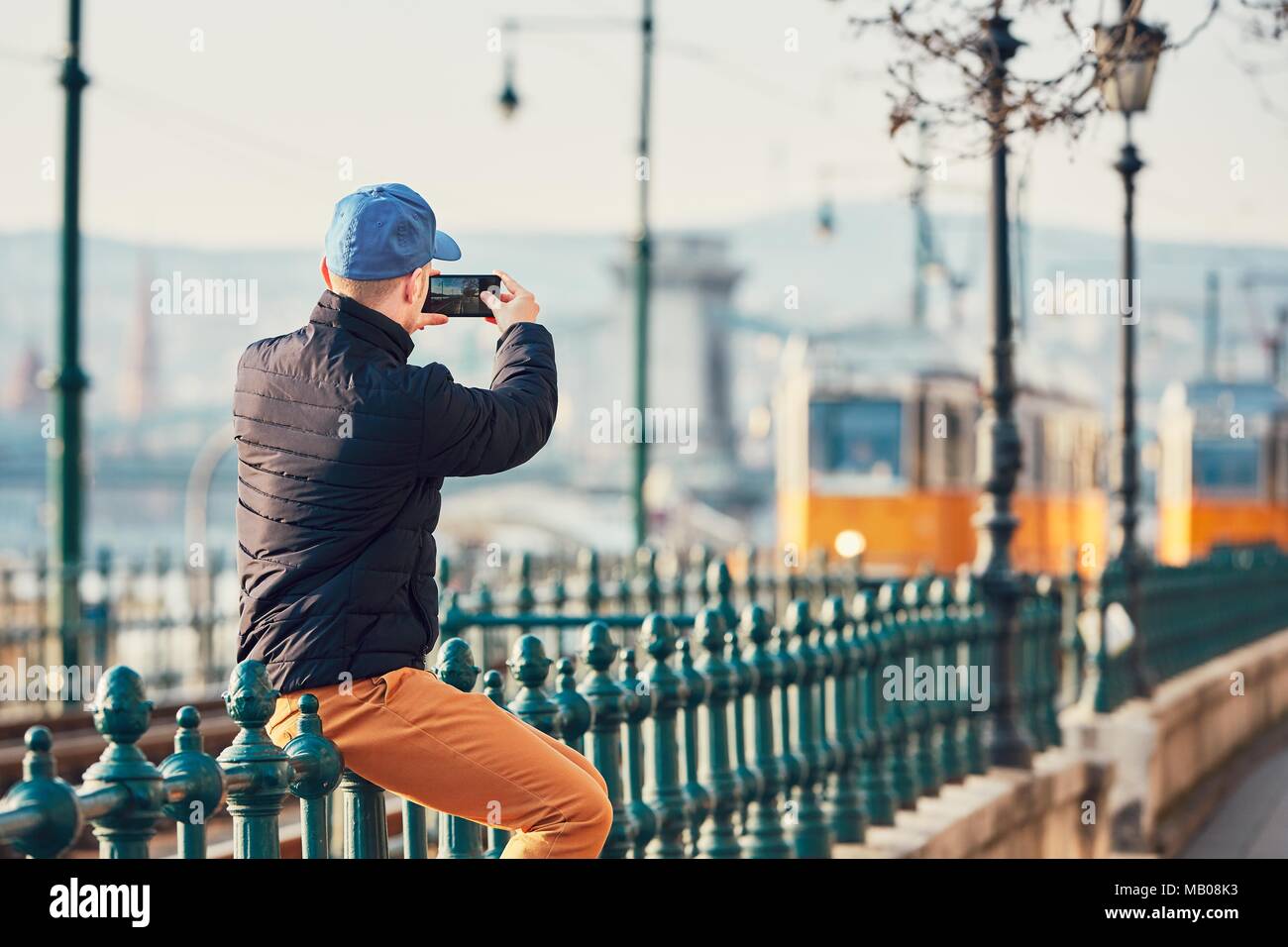 Jeune homme (touriste) homme assis sur la rambarde et prendre photo avec téléphone intelligent. Les Trams à Budapest, Hongrie. Banque D'Images