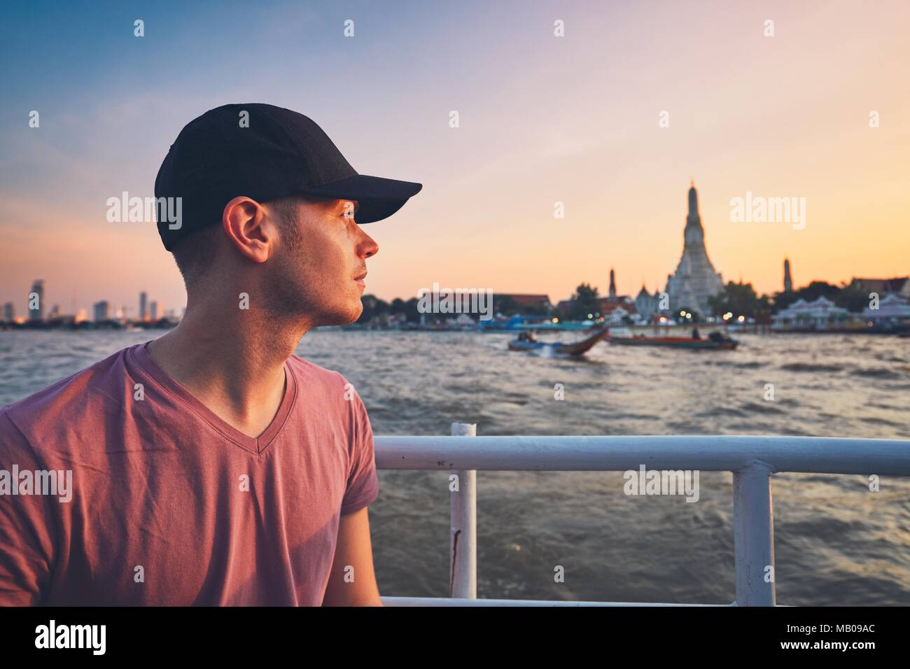 Jeune homme avec bouchon sur le bateau contre temple Wat Arun au coucher du soleil. Chao Phraya à Bangkok, Thaïlande. Banque D'Images