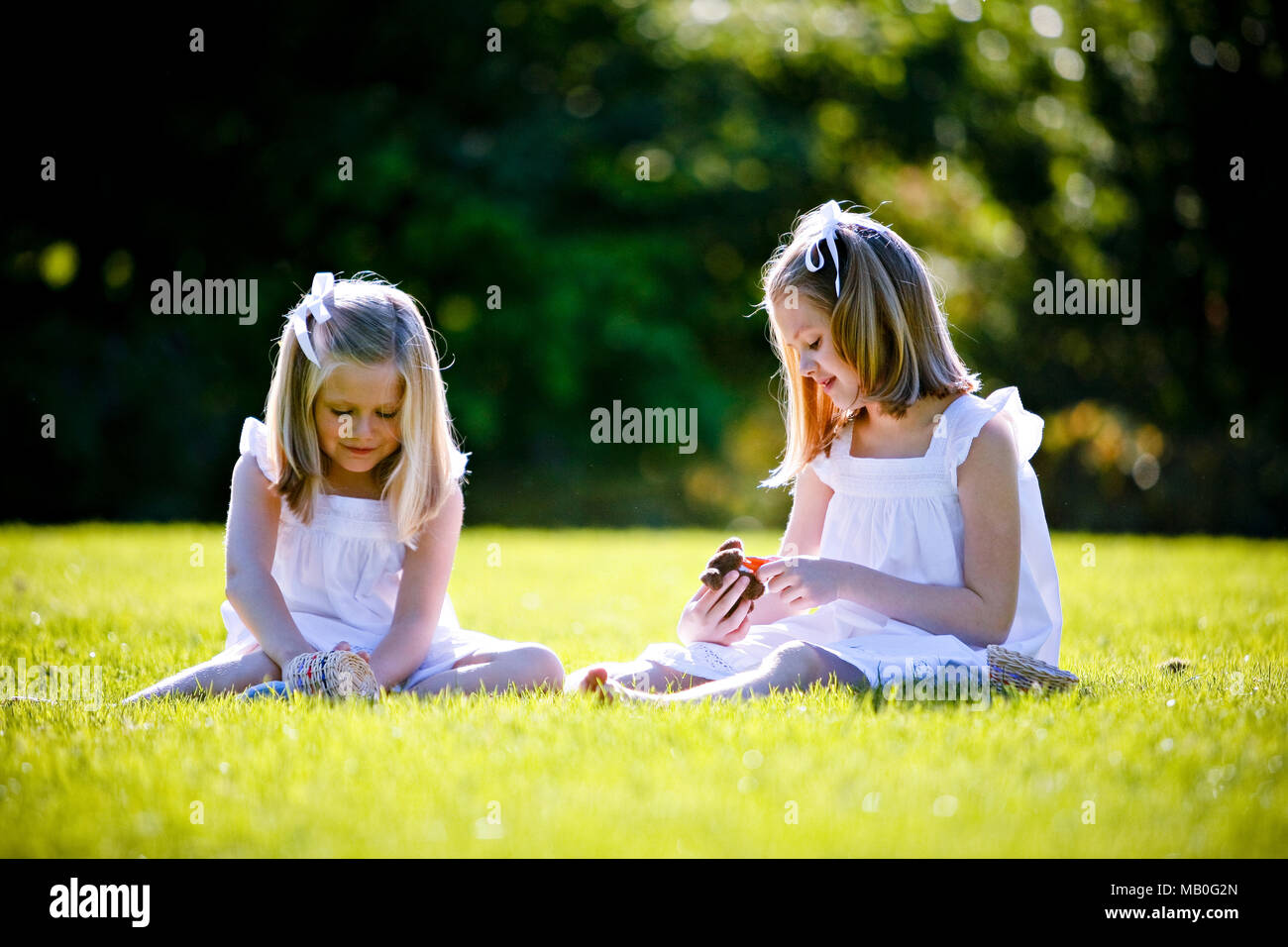 Deux jeunes très Caucansian in white jupes sitting on grass jouant à un parc avec éclairage arrière et trouble fond vert Banque D'Images