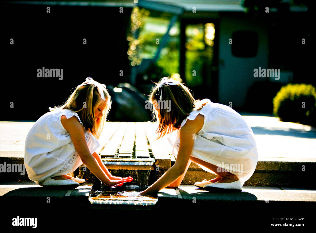 Deux jolies jeunes filles en jupes blanches et chaussures blanches jouant avec l'eau de la fontaine dans un parc avec la lumière du soleil qui brille de derrière Banque D'Images