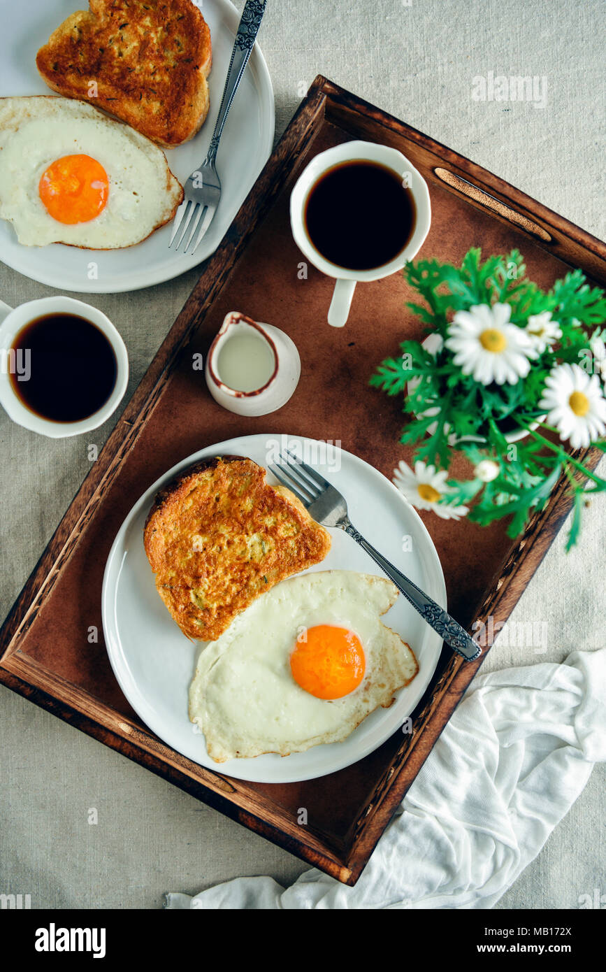 Easy French Toast servi avec un œuf frit sur le côté sur une plaque blanche, une tasse de café, le lait et les fleurs de printemps dans un plateau en bois photographié de à Banque D'Images