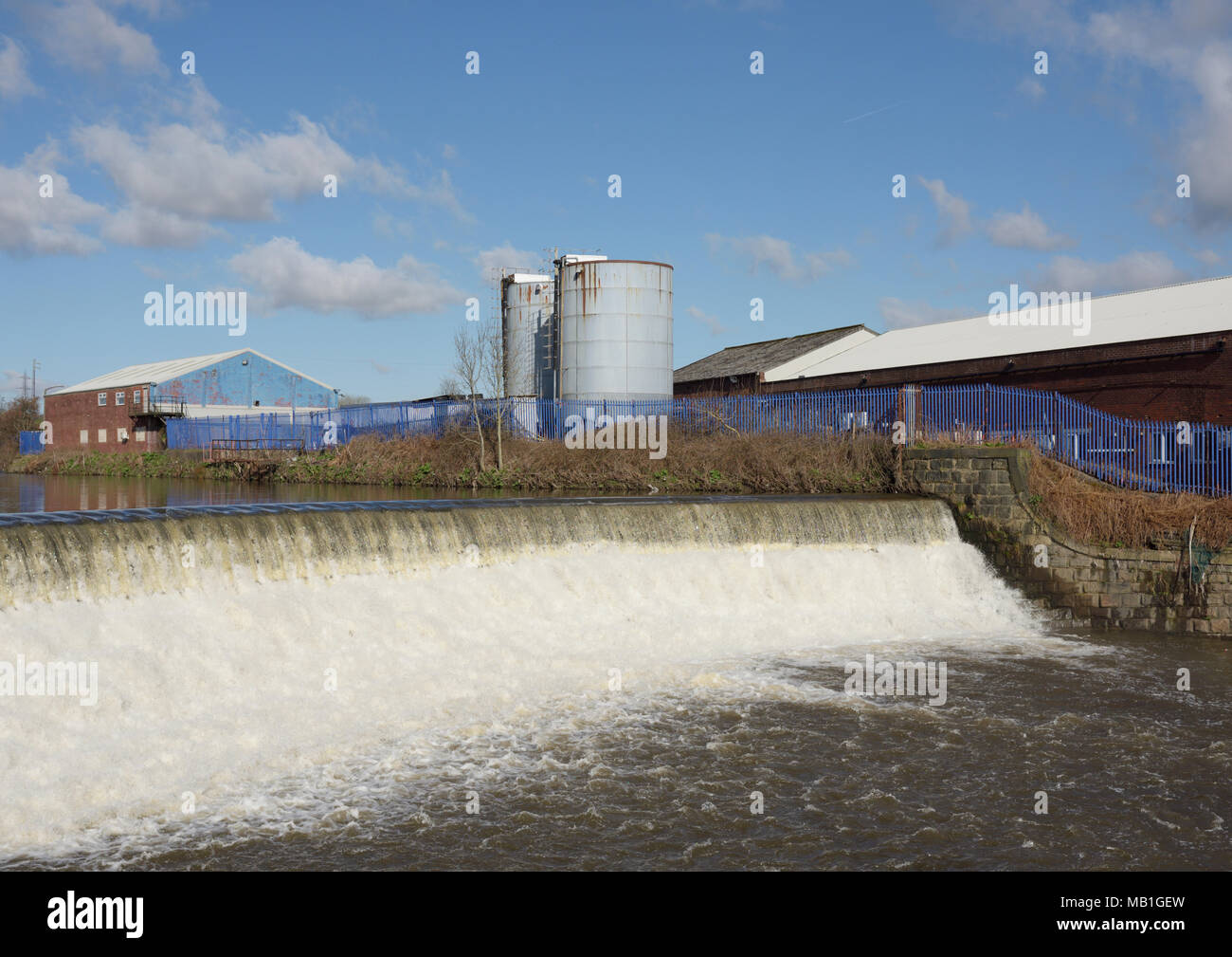 Weir sur la rivière irwell provoquant un écoulement turbulent d'eau en aval, deux silos bleus et des bâtiments industriels sur la rive, radcliffe bury lancashire royaume-uni Banque D'Images