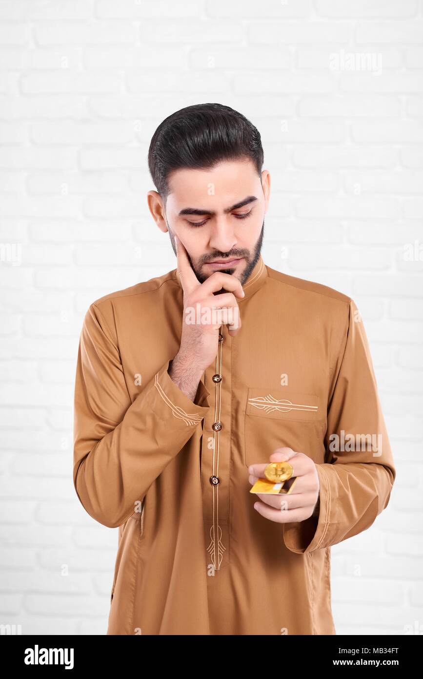 Un homme musulman sérieux et bitcoin permet une carte de crédit. Ressemble à ce qu'il doit prendre une décision importante. Il porte une chemise beige avec un motif. Photo a été fait sur fond blanc studio. Banque D'Images