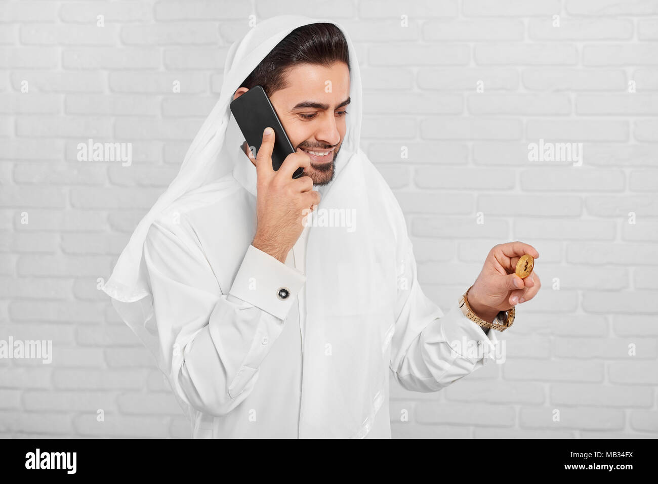Un musulman businessman wearing costume traditionnel blanc, regarde sur bitcoin doré dans sa main. Dans un autre côté, il garde son téléphone portable noir. Il porte golden watch et boutons de manchette sur sa chemise. Banque D'Images