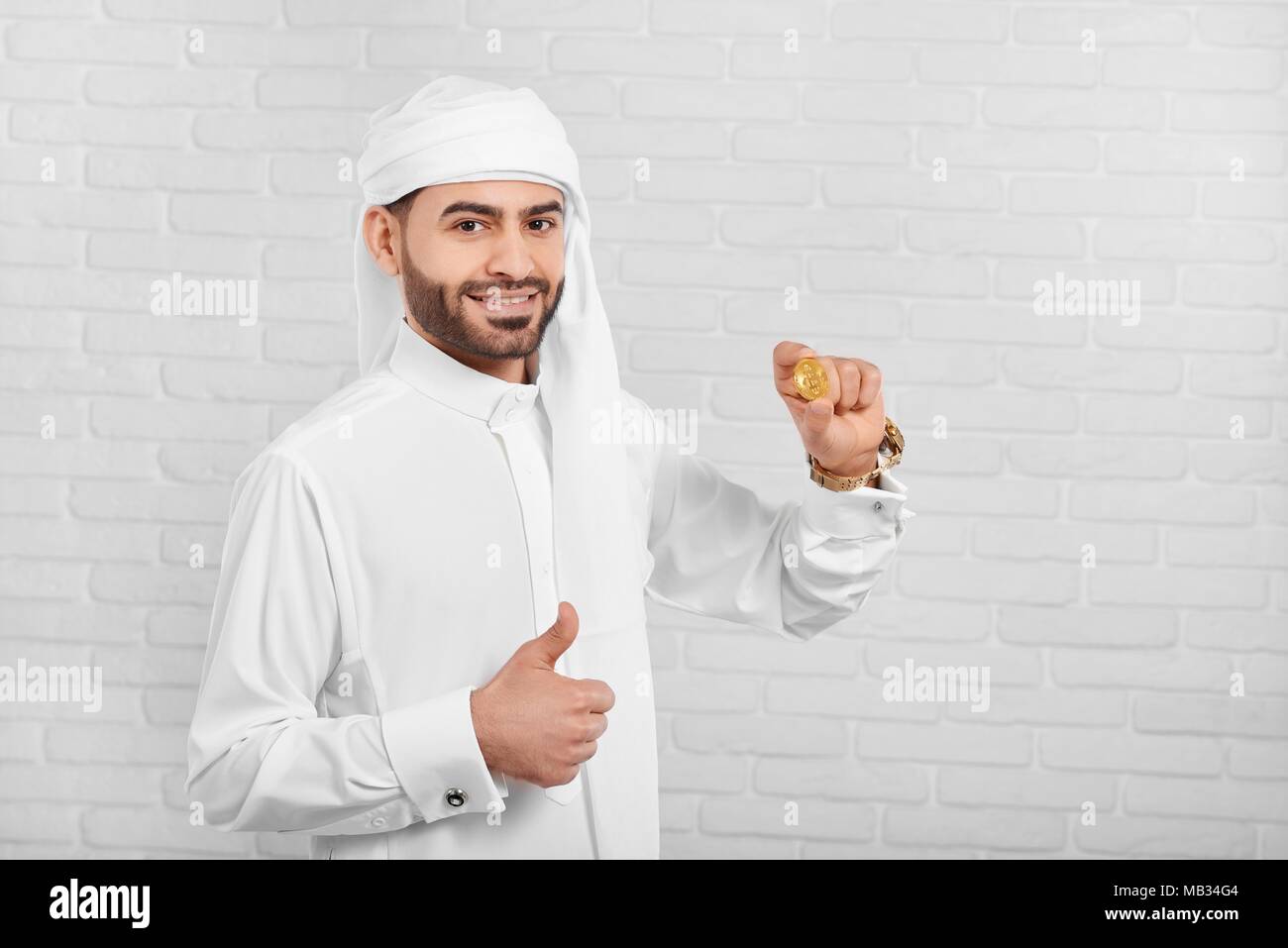 L'homme conserve arabe souriante et bitcoin semble très heureux. Il porte le costume traditionnel arabe blanc,golden watch et les boutons de manchette. Un homme d'affaires semble très positive et heureuse. Banque D'Images