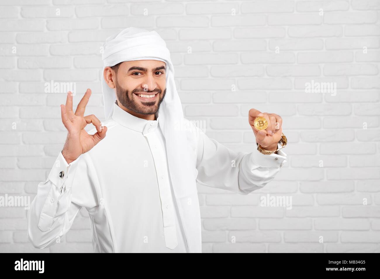 L'homme conserve arabe souriante et a l'air très heureux des bitcoins, debout sur fond blanc. Il porte le costume traditionnel arabe blanc,golden watch et les boutons de manchette. Un homme d'affaires semble positive et heureuse. Banque D'Images