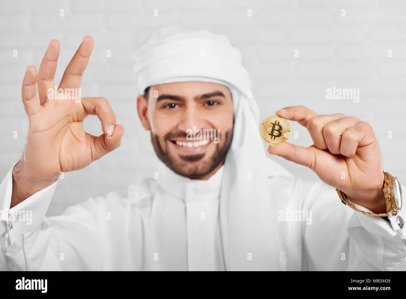 Studio Portrait of a smiling man arabe et bitcoin permet a l'air très heureux, debout sur fond blanc. Il porte le costume traditionnel arabe blanc,golden watch et les boutons de manchette. Banque D'Images
