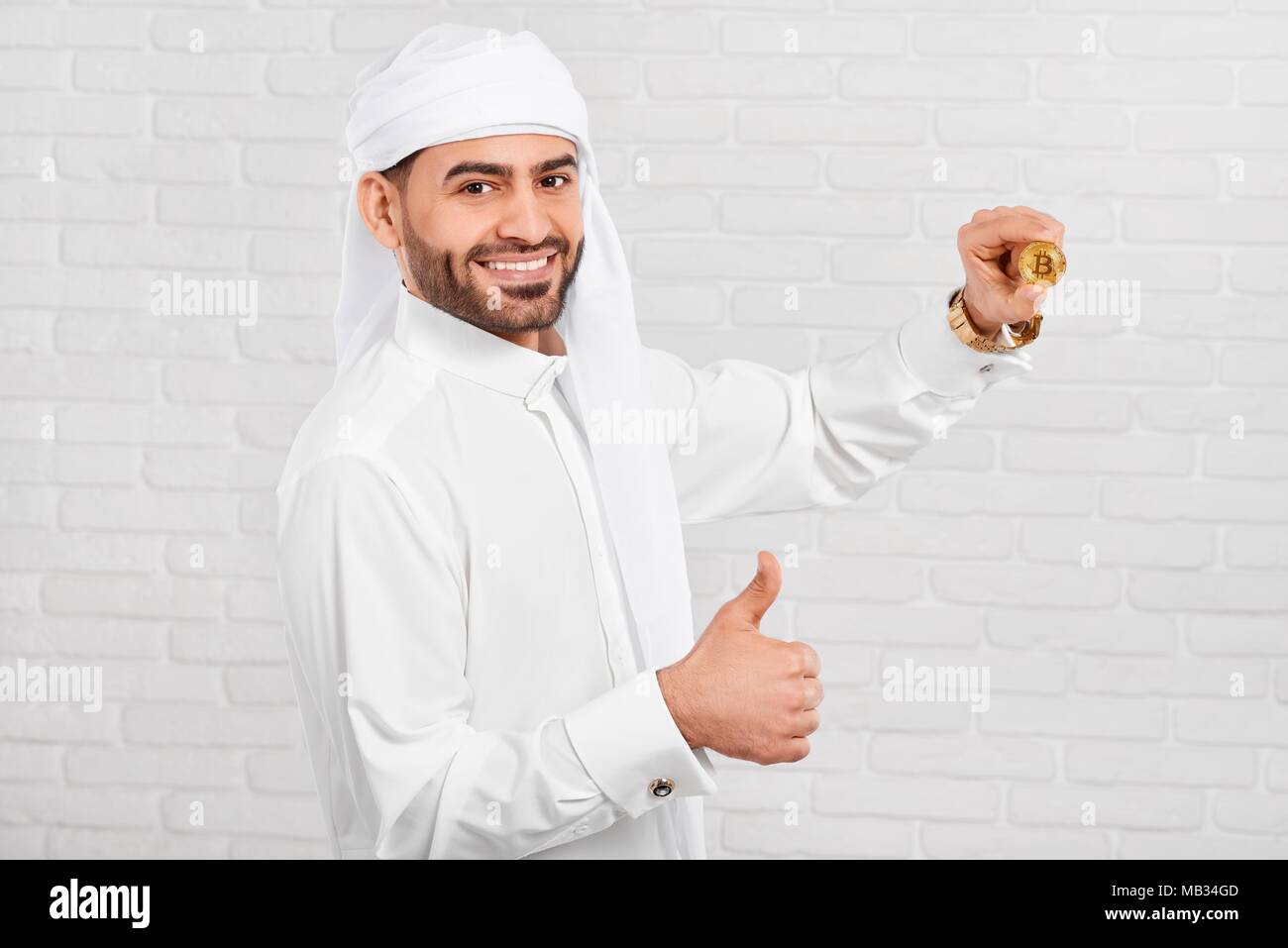 Studio photo of a smiling woman musulmane et bitcoin permet a l'air très heureux, debout sur fond blanc. Il porte le costume traditionnel arabe blanc,golden watch et les boutons de manchette. Banque D'Images