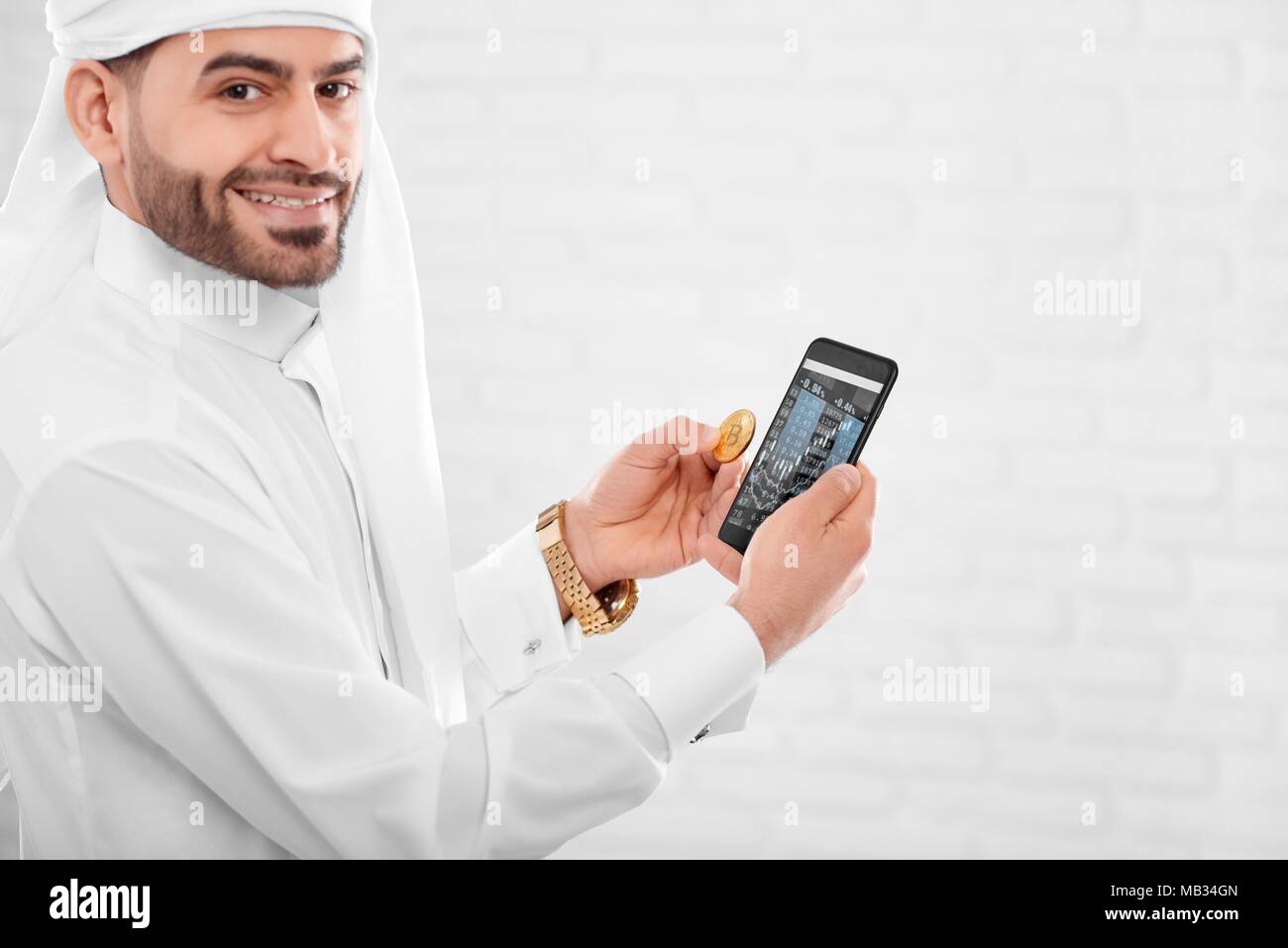 Jeune musulman homme conserve bitcoin or et du téléphone mobile dans une autre main. Il est sur le fond de studio blanc. Homme d'un air heureux et engagé. Banque D'Images