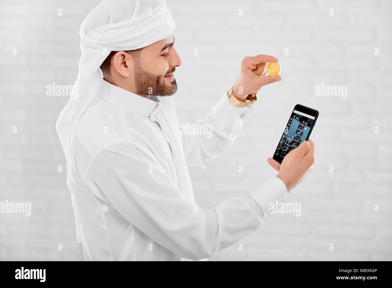 Jeune musulman maintient l'opérateur de téléphonie mobile et pièce d'or dans l'autre main. Il est sur le fond de studio blanc. Un homme a l'air heureux et impliqués. Banque D'Images