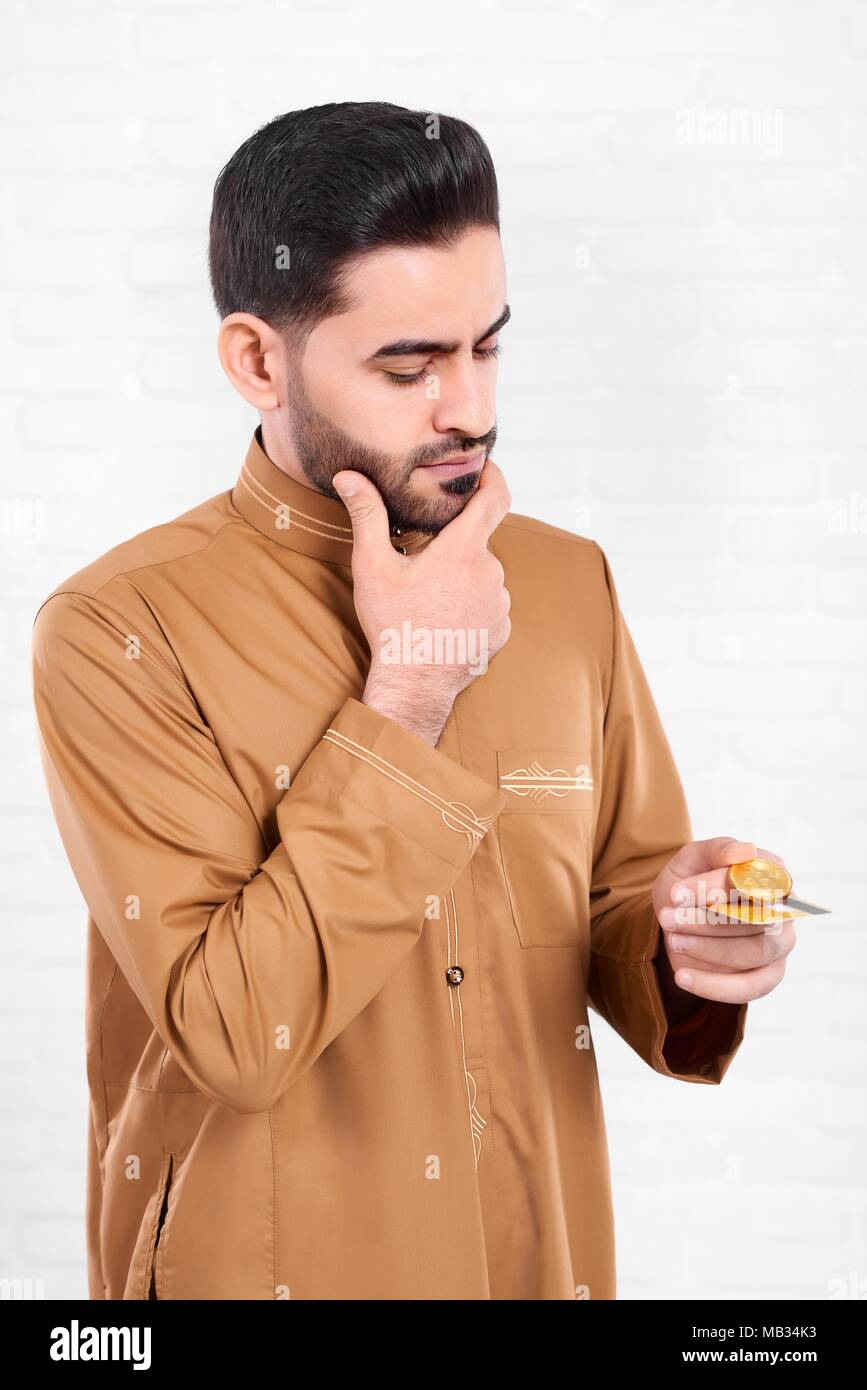 Un homme noir avec entendre , porter chemise beige avec le modèle précis, se dresse sur le fond blanc de studio. Il est réfléchi et garde dans ses mains - pièce d'or et bitcoin une carte de crédit. Banque D'Images