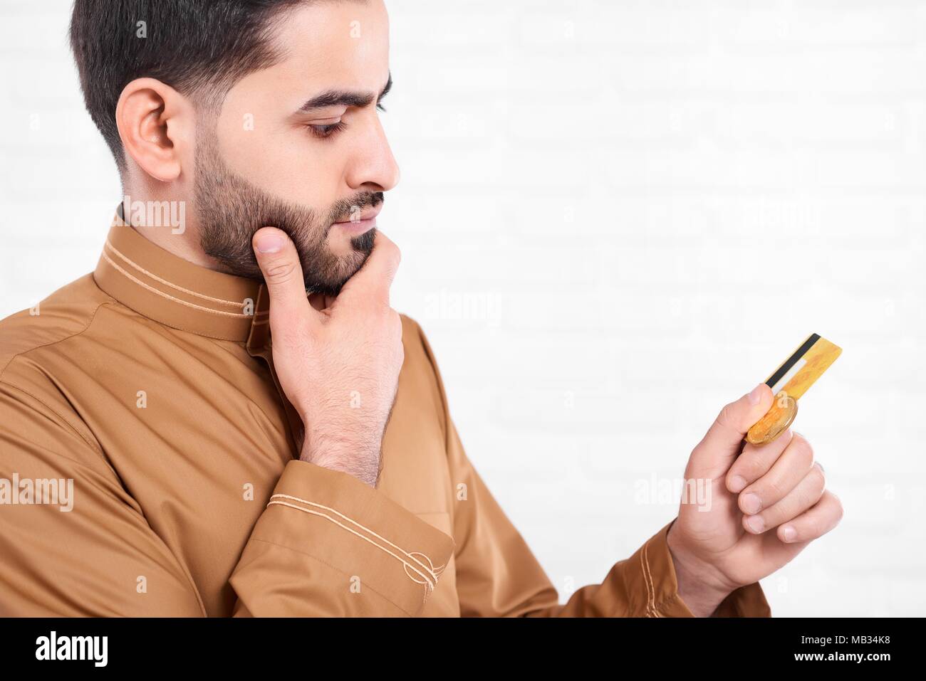 Un musulman noir avec entendre , porter chemise beige avec le modèle précis, se dresse sur le fond blanc de studio. Il tient dans ses mains des bitcoins en or et une carte de crédit. Banque D'Images
