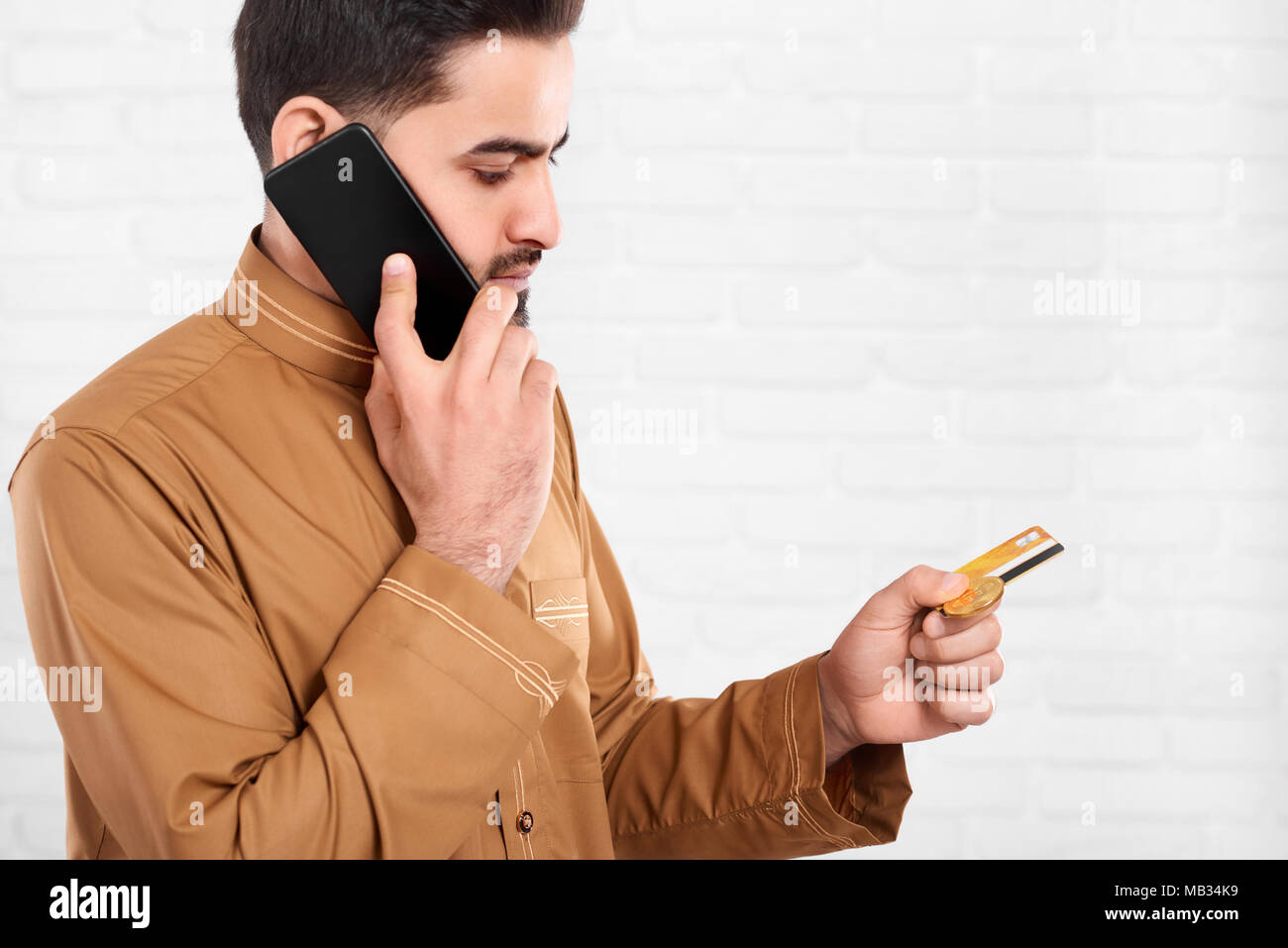 Le jeune homme d'Oman continue de carte de crédit et parle au téléphone. Il a un grand bitcoin d'or dans ses mains. Il porte la chemise beige avec un motif. Un homme se tient sur le fond blanc de studio. Banque D'Images