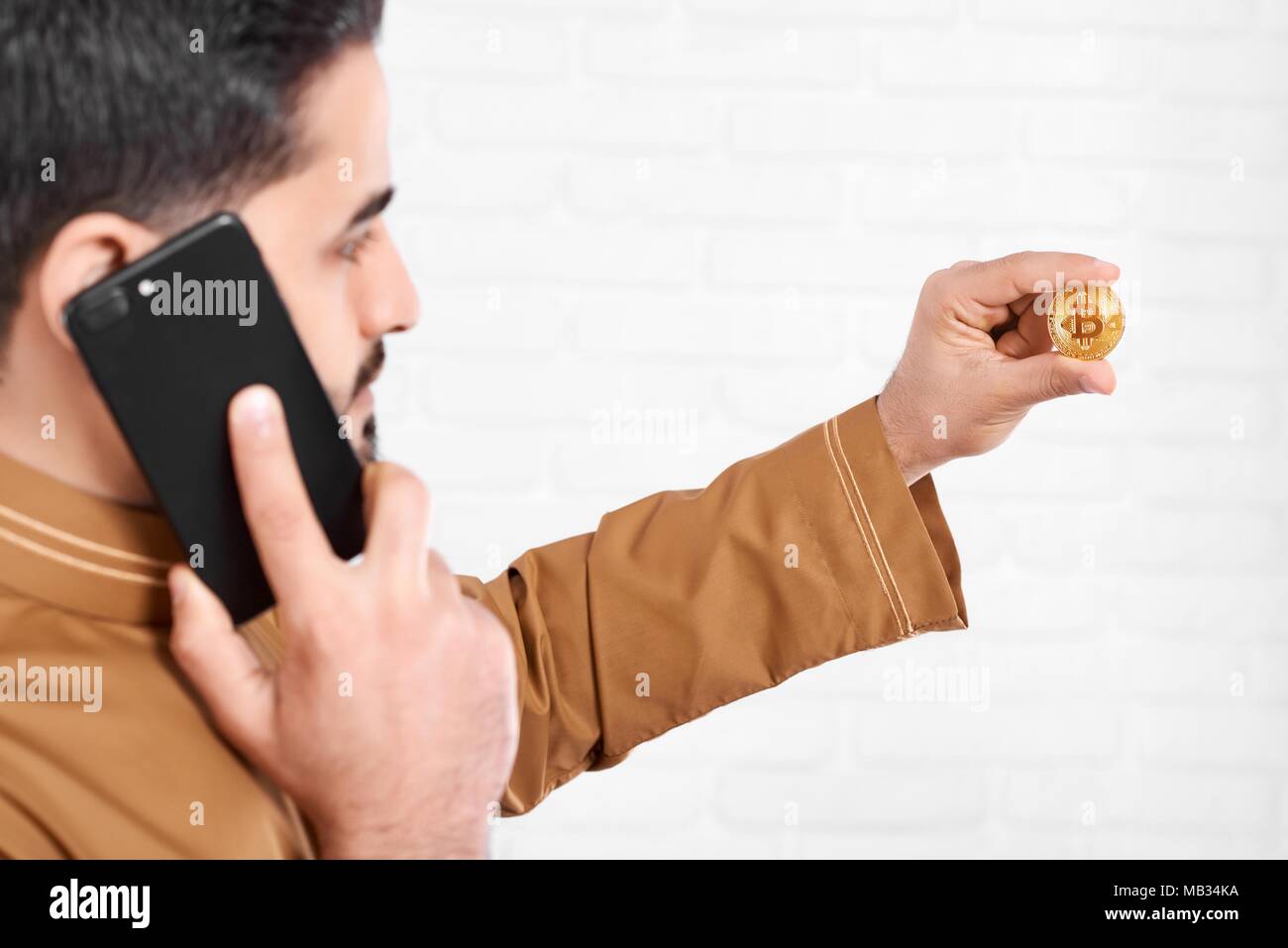 Un jeune homme conserve un brillant or bitcoin. Un homme parle sur son IPhone noir et regarde sur la pièce dans sa main. Il est sur le fond de studio blanc. Banque D'Images