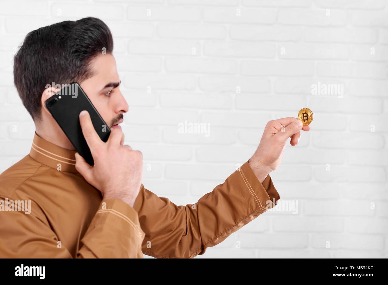 Un jeune homme d'attrayants conserve un brillant or bitcoin. Un homme parle sur son téléphone mobile noir et regarde sur la pièce dans sa main. Il est sur le fond de studio blanc. Banque D'Images