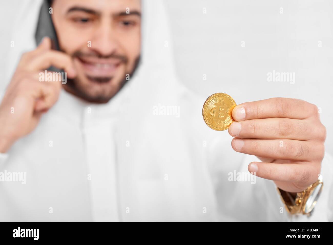 Un musulman businessman wearing costume traditionnel blanc, regarde sur bitcoin doré dans sa main. Dans un autre côté, il garde son téléphone portable noir. Il porte golden watch et l'air heureux. Banque D'Images