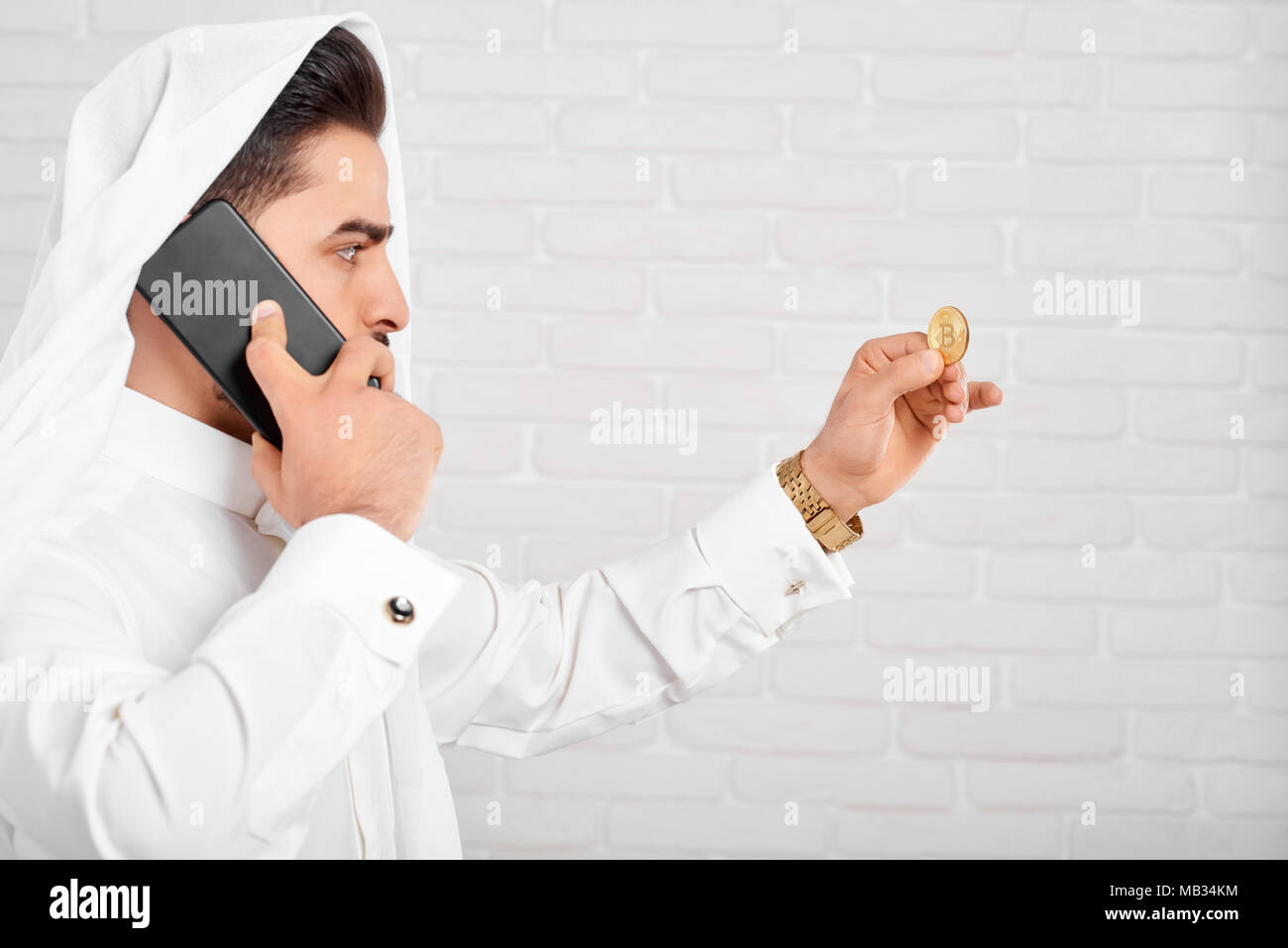 Un homme d'affaires arabe portant costume traditionnel blanc, regarde sur bitcoin doré dans sa main. Dans un autre côté, il garde son téléphone mobile noir et de pourparlers. Close-up faite sur un arrière-plan blanc studio. Banque D'Images