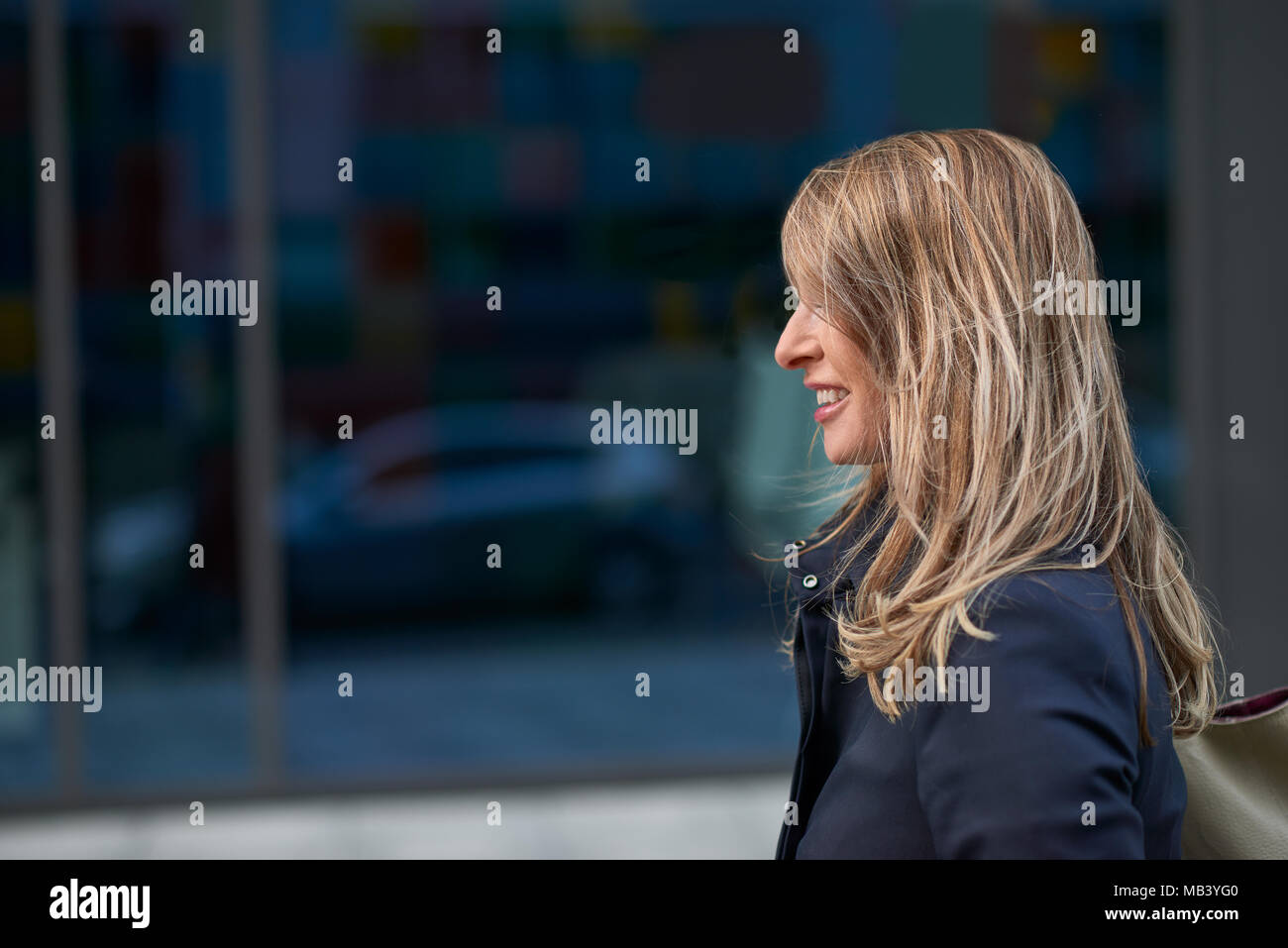 Happy blonde femme avec cheveux ébouriffé par le vent marchant dans une rue urbaine passant une vitrine commerciale avec la réflexion d'une automobile et de copie Banque D'Images