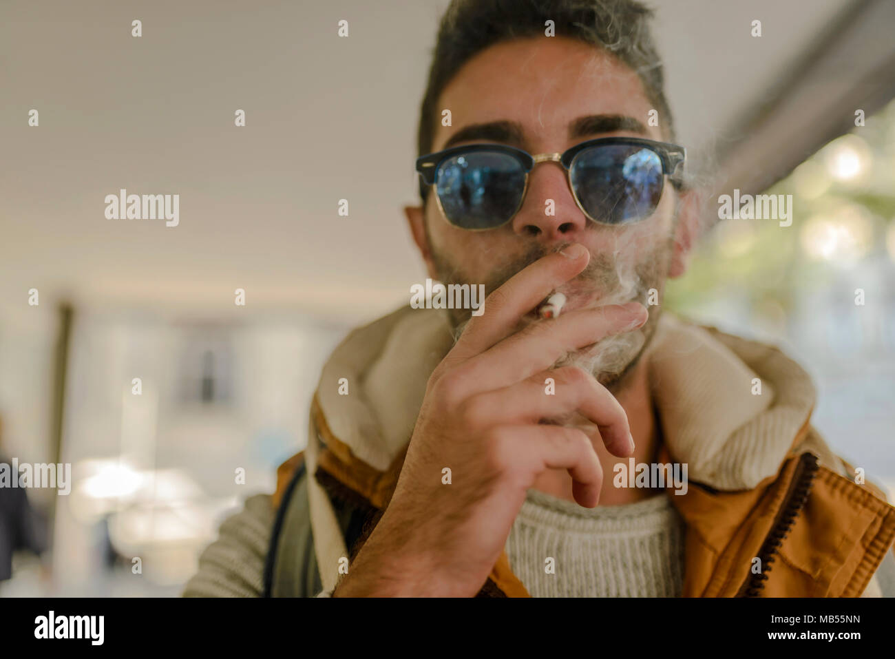 Beau jeune hipster avec orange jacket and sunglasses bénéficiant d'une cigarette dans la rue. Banque D'Images