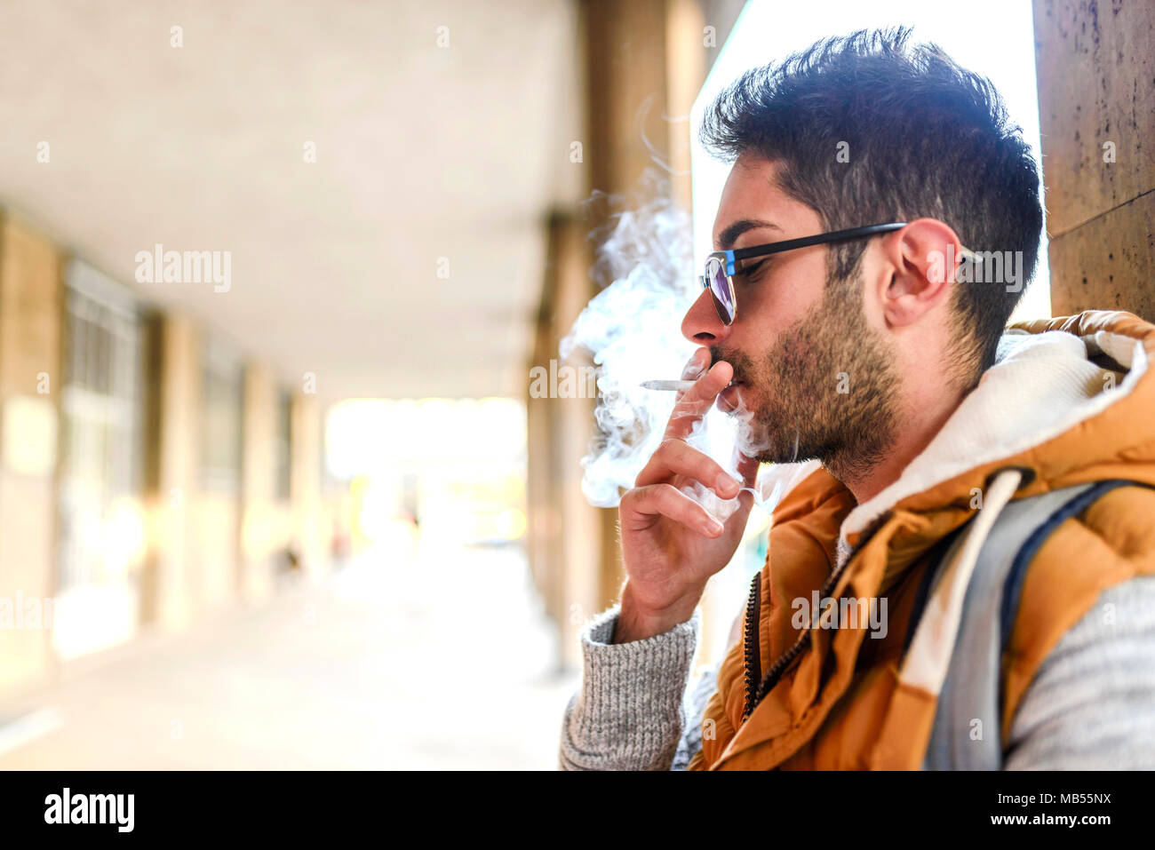 Beau jeune hipster avec orange jacket and sunglasses bénéficiant d'une cigarette dans la rue. Banque D'Images