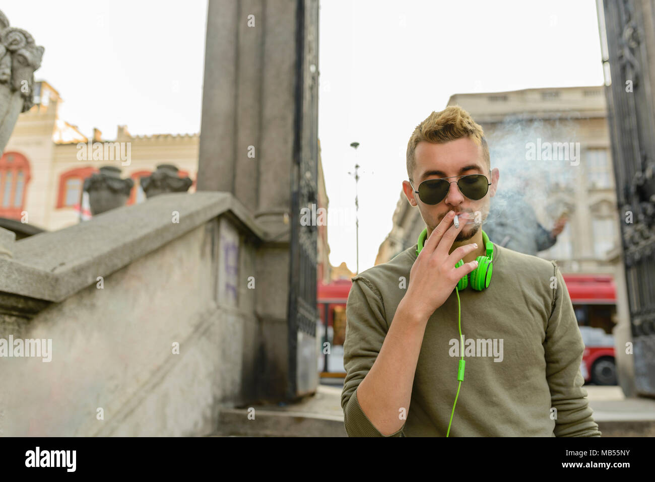 Jeune homme séduisant avec casque vert sunglasse et bénéficiant d'une cigarette dans la rue. Banque D'Images