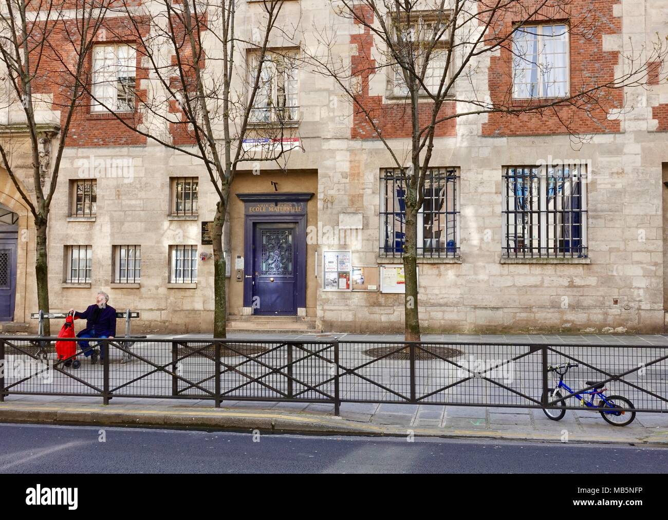 Homme avec un panier rouge est assis sur un banc en face de l'école maternelle, l'école maternelle française, 4e arrondissement, Paris, France Banque D'Images