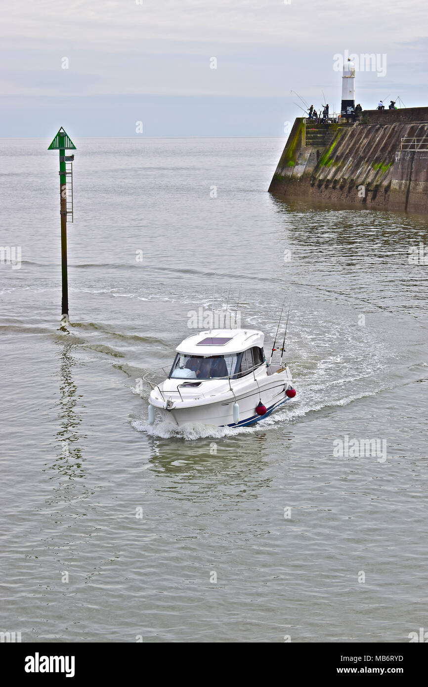 Un petit bateau retourne à la sécurité de Porthcawl Marina après une journée de pêche dans le canal de Bristol avec les pêcheurs sur le mur du port derrière. Banque D'Images