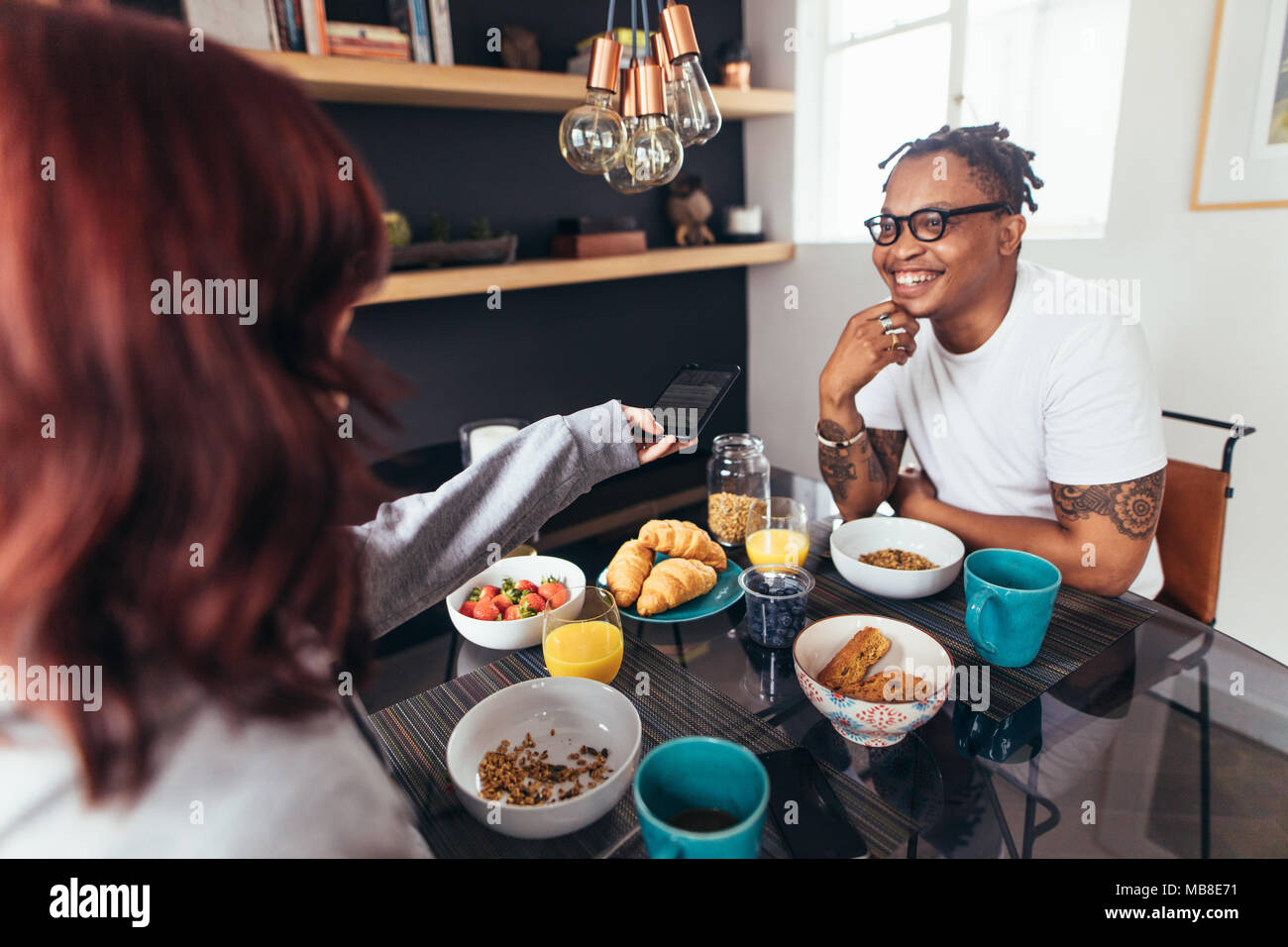 Happy young couple having breakfast ensemble à la maison. Woman giving téléphone mobile pour son petit ami souriant assis à table de petit déjeuner. Banque D'Images