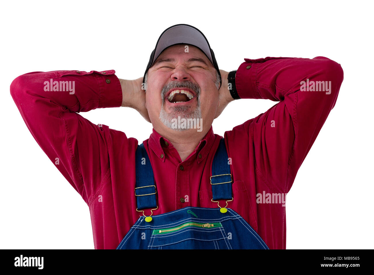 Agriculteur jovial ou travailleur dans le chapeau et salopette denim bénéficiant d'un rire chaleureux comme il jette la tête en arrière, avec les mains jointes derrière son cou isolé sur w Banque D'Images