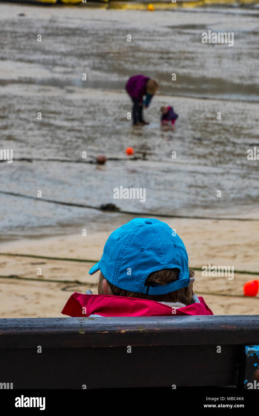 Un ancien ou un homme âgé assis au bord de la mer, sur un banc avec vue sur le port de personnes jouant sur la plage. Bleu portant une casquette de baseball. Banque D'Images