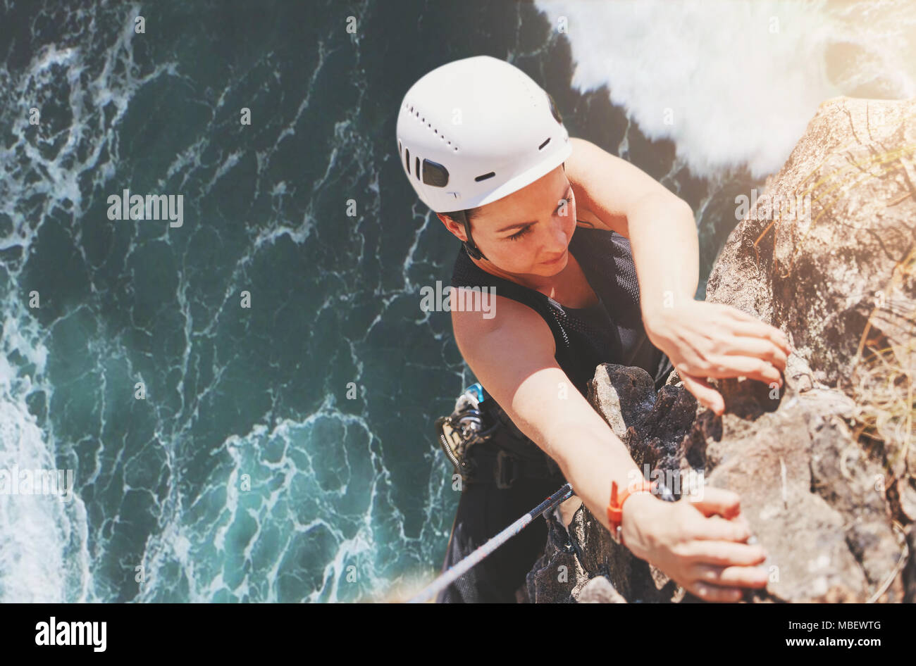 Déterminé, l'accent female rock climber hanging from rock au-dessus de l'océan ensoleillé Banque D'Images