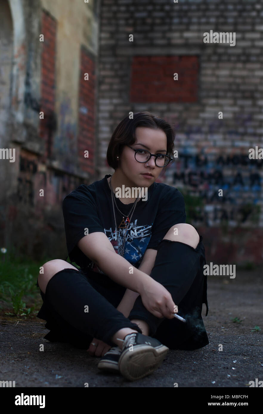 Une fille avec de courts cheveux foncés, lunettes, My Chemical Romance un t-shirt et d'une cigarette assis près d'un bâtiment ancien. Banque D'Images