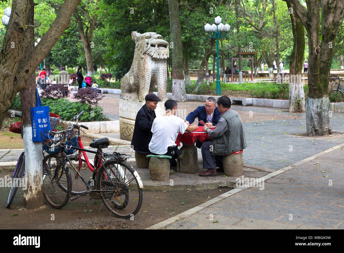 Quatre hommes chinois s'asseoir cartes à jouer dans le parc de Yangshuo, Chine. Leurs bicyclettes reste contre un arbre. Derrière un lion gardien chinois pierre monte la garde. Banque D'Images