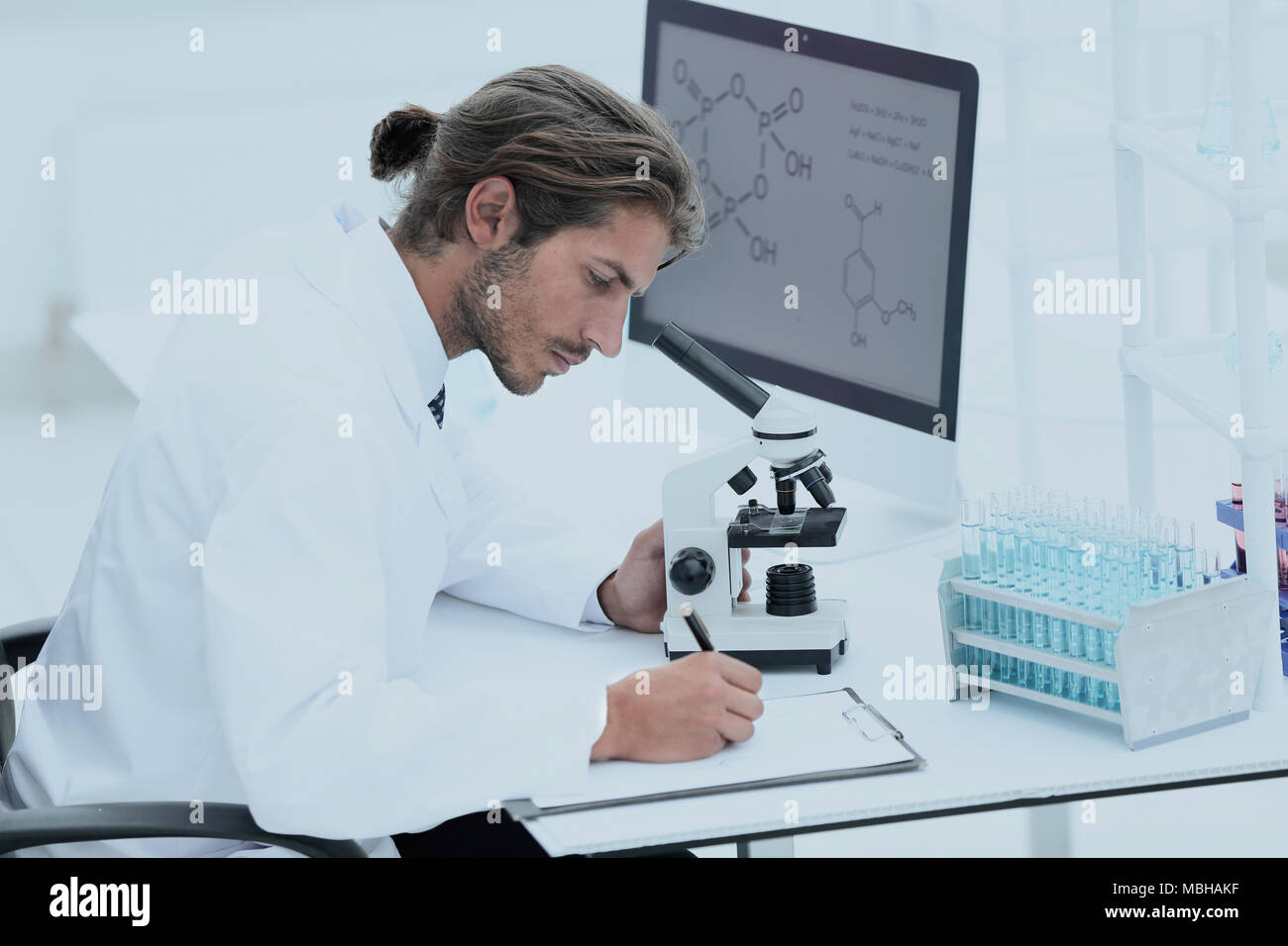 Un jeune chercheur ou spécialiste technique prend des notes dans le laboratoire d'histopathologie en utilisant un microscope optique et un ordinateur Banque D'Images