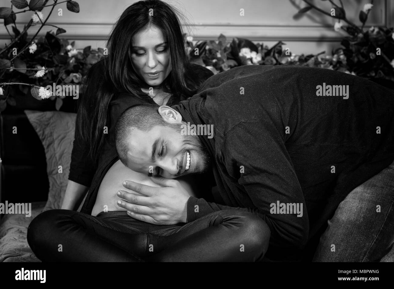 Photo en noir et blanc d'enamouré couple international, l'homme écoute le ventre de sa femme enceinte, assise sur le lit douillet gris dans la chambre à coucher Banque D'Images