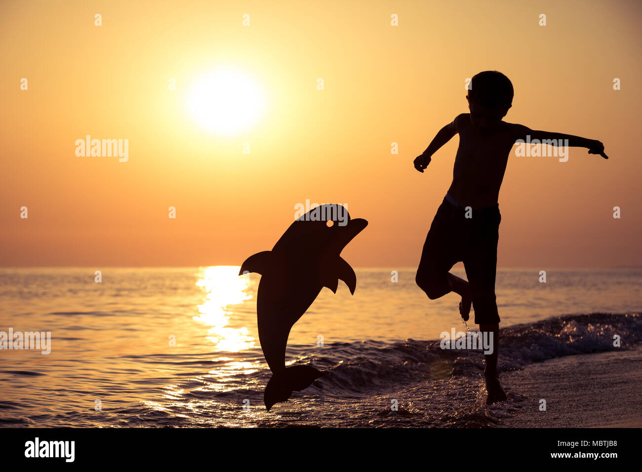 Un petit garçon heureux d'exécution sur la plage, à l'heure du coucher du  soleil. Il joue avec un dauphin en carton. Kid s'amusant à l'extérieur.  Concept de vacatio d'été Photo Stock -