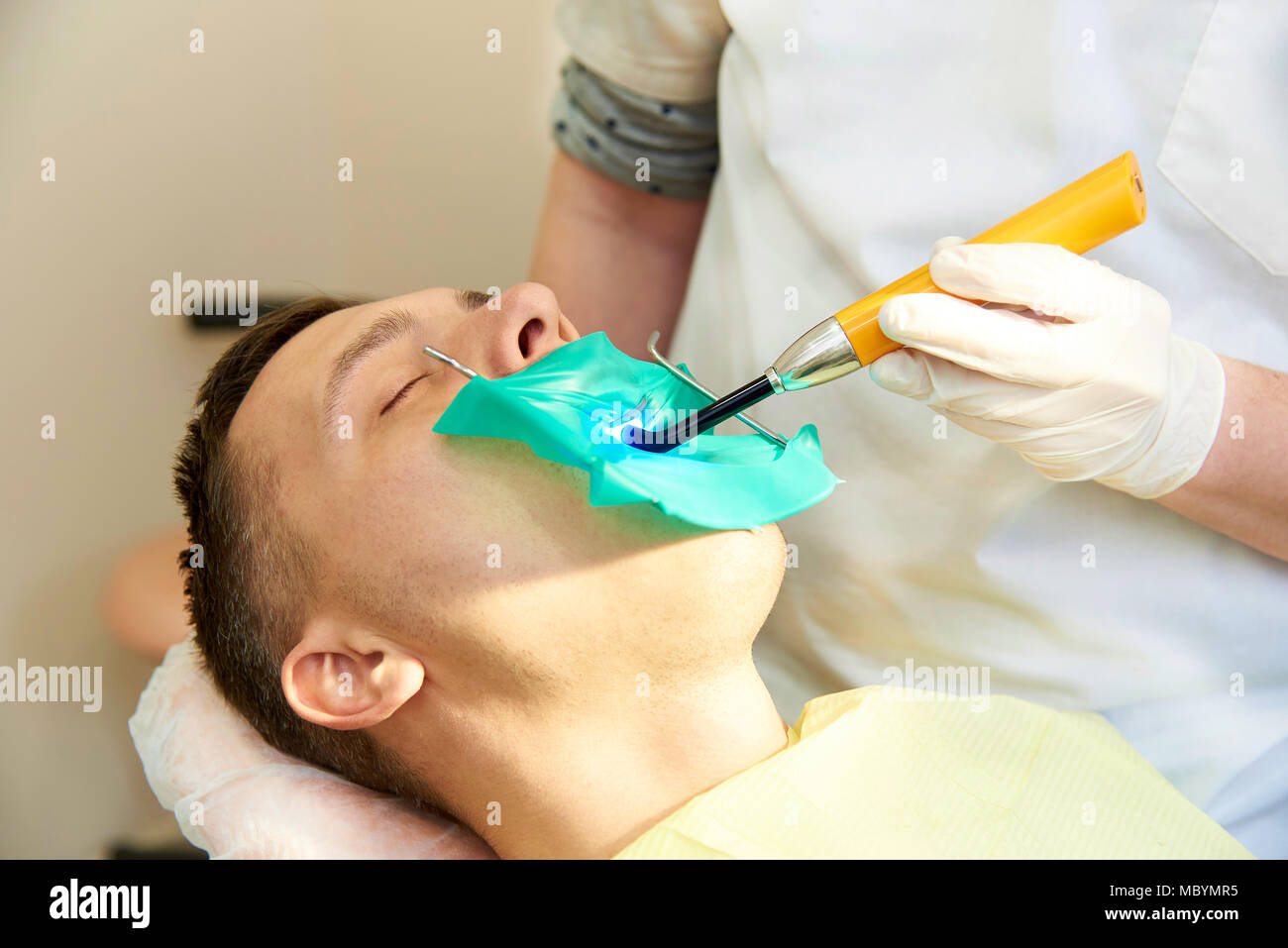 Un jeune homme aux yeux clos sur le fauteuil dentaire. Le dentiste travaille avec une lampe de polymérisation dentaire dans la bouche du patient. Banque D'Images