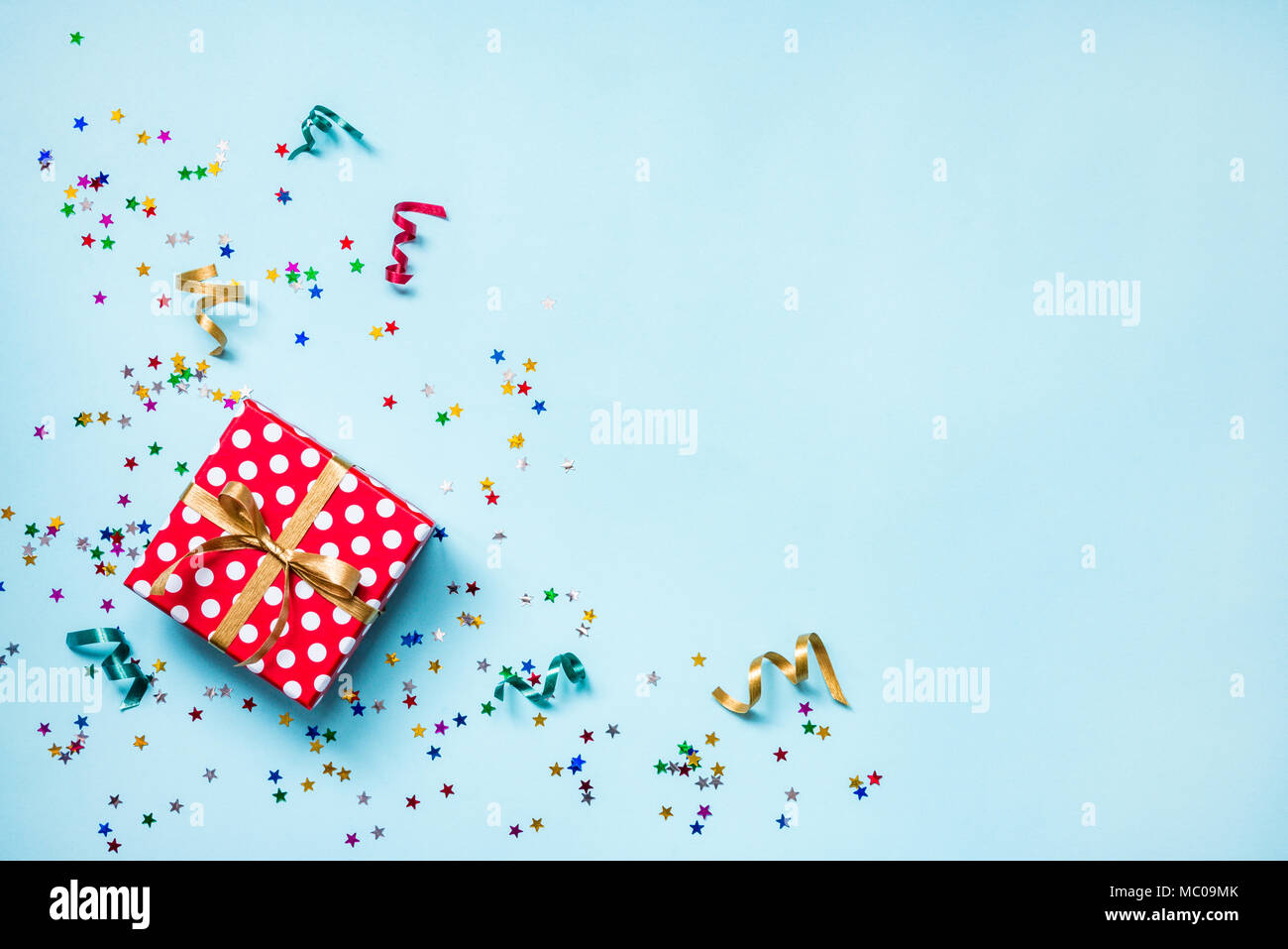 Vue de dessus d'une boîte cadeau rouge en pointillés, dispersés en forme de confettis étoile scintillante et rubans colorés sur fond bleu. Concept de célébration. Savs copie Banque D'Images