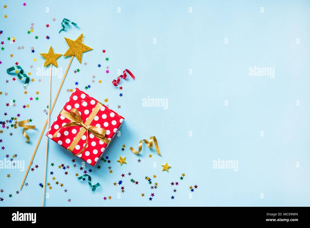 Vue de dessus d'une boîte cadeau rouge en pointillés, golden baguettes magiques, confetti colorés et rubans sur fond bleu. Concept de célébration. Copier l'espace. Banque D'Images