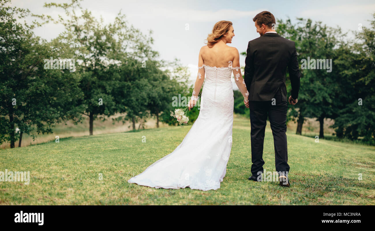 Vue arrière de la belle jeune femme dans une robe de mariage qui tient la main de l'époux et la marche dans le parc. Couple holding hands and walking outdoors sur Banque D'Images Vue arrière de la belle jeune femme dans une robe de mariage qui tient la main de l'époux et la marche dans le parc. Couple holding hands and walking outdoors sur Banque D'Images