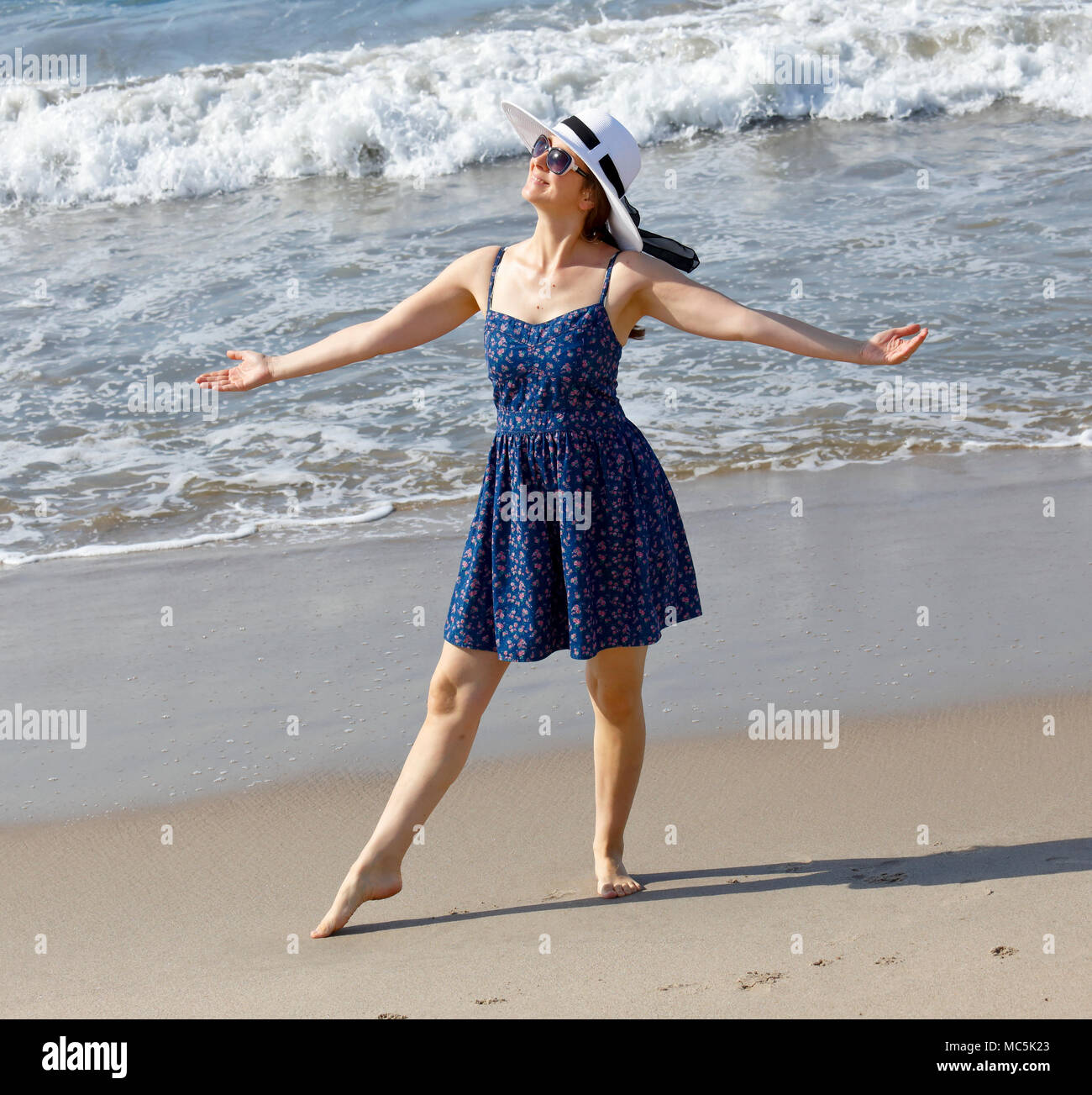 Jeune femme sur une plage dans une joyeuse posent avec bras ouverts. Il porte une robe bleue, un chapeau blanc, et des lunettes de soleil Banque D'Images