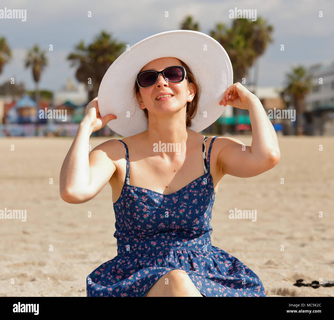 Jeune femme en blanc chapeau, lunettes de soleil, et bleu robe d'été. Elle est maintenant le bord de son chapeau et a sa tête légèrement inclinée vers le haut Banque D'Images