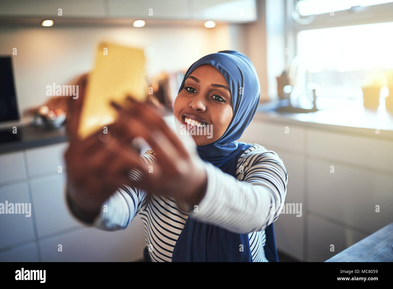 Souriante jeune femme arabe portant un hijab en tenant vos autoportraits avec son portable alors qu'il était assis dans sa cuisine à la maison Banque D'Images