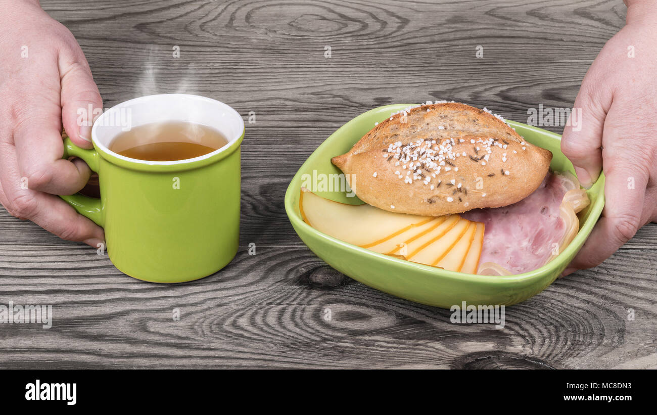 Les mains sont au service de petit déjeuner avec du thé chaud. Bol vert, salé pain, tranches de jarret et d'edam, mug sur le fumeur verre sur fond de bois. Banque D'Images
